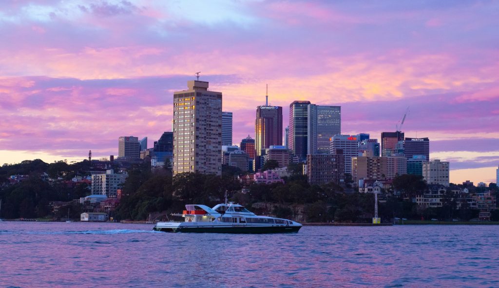 A Sydney fast ferry on the water with North Sydney in the background at sunset, with the sky glowing purple.